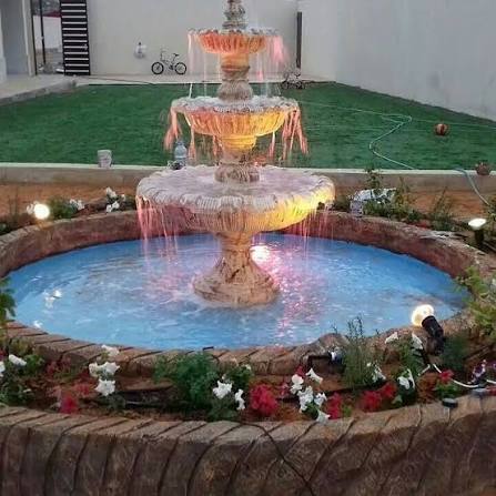 نافورة حجرية متدرجة داخل حوض دائري بإنارة ملونة وزهور محيطة